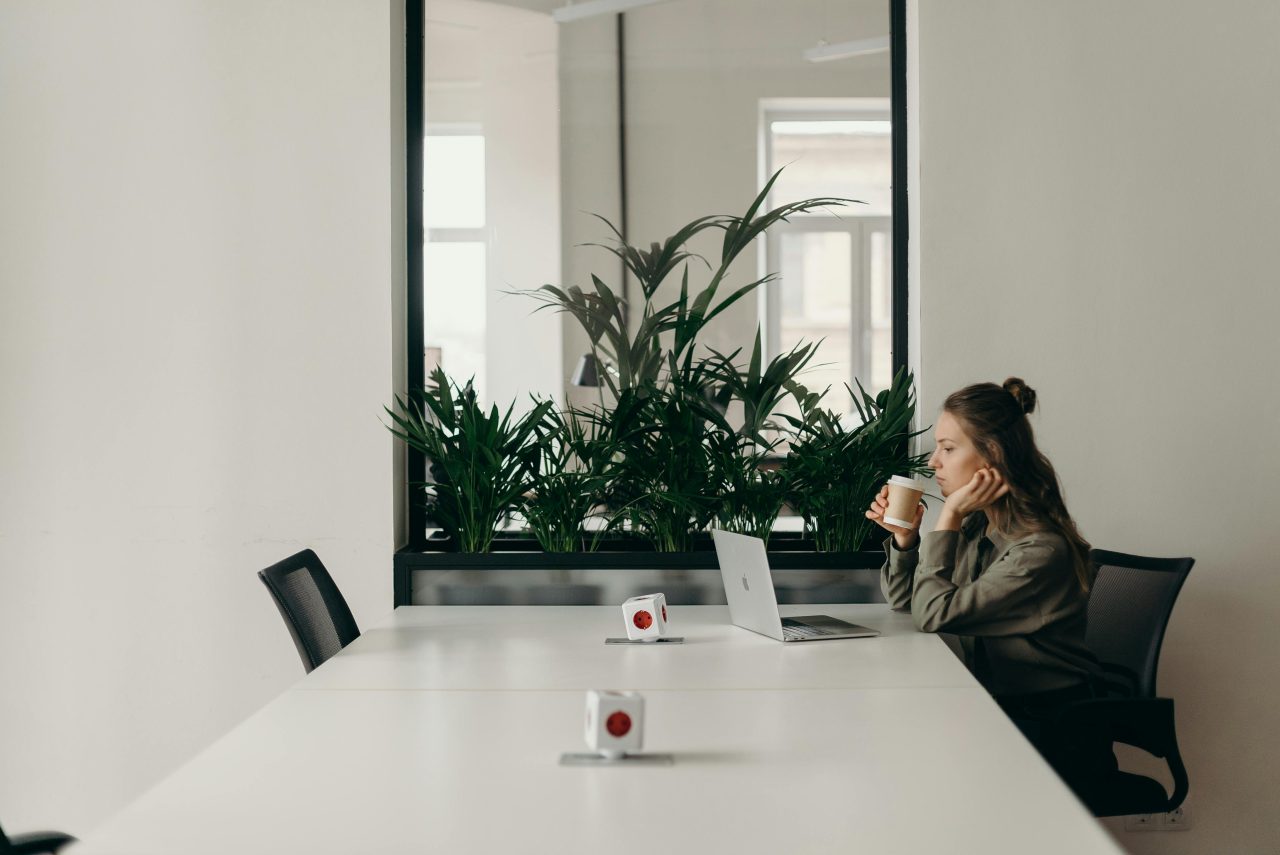 A woman sits at a glossy office table with empty seats around her while she sips a take-out coffee and looks at her laptop