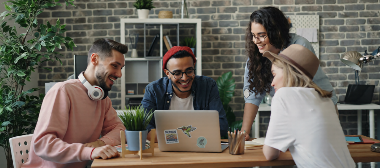Four coworkers sit at a table looking at a laptop together. They are happy and enjoying the interaction